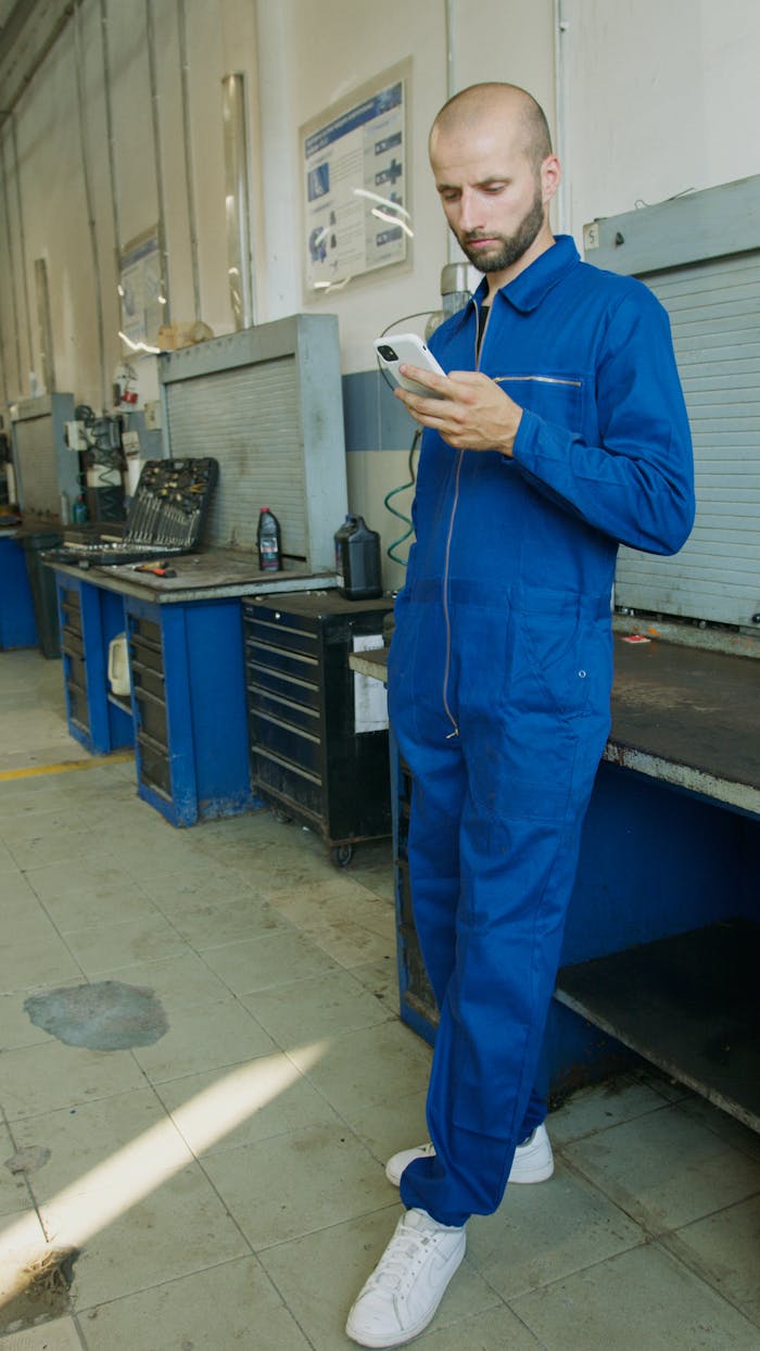 about-03 A bald mechanic in a blue jumpsuit checks his smartphone in an industrial workshop setting.